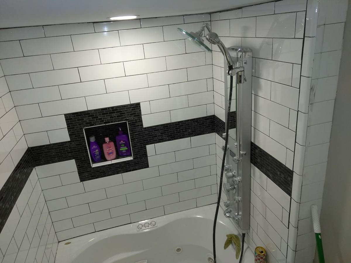 The same tub and shower tiled in white subway tile with a black mosaic band running around it and a finished niche holding three bottles.