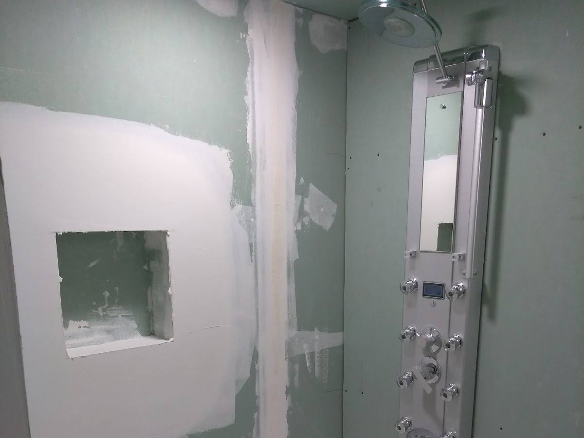 A tub and shower stall with bare green drywall, taped seams not yet sanded, a rough cut-out where a shelf niche will go, and the shower panel already mounted.