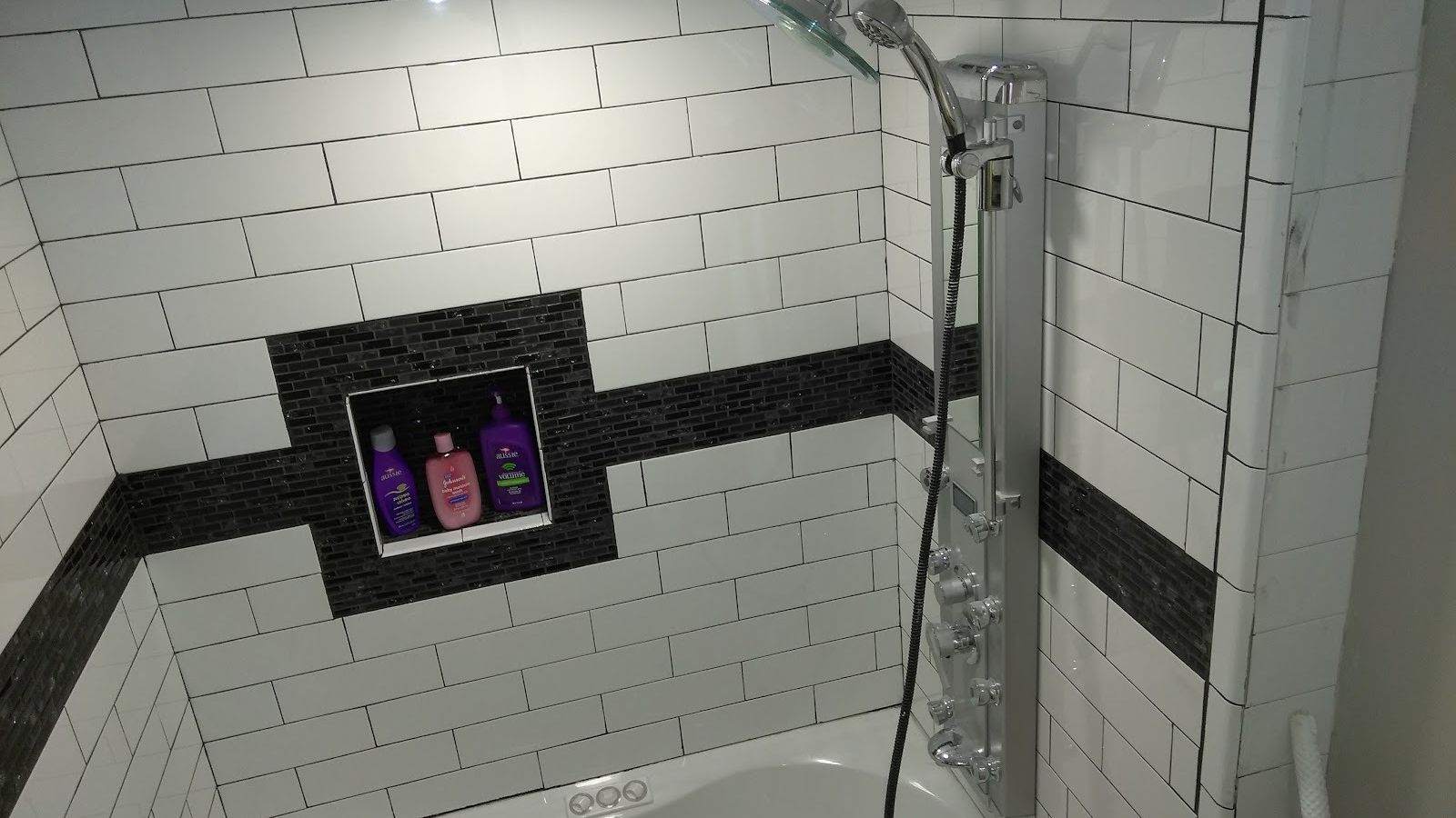 Floor-to-ceiling white subway tile around a tub and shower, with a black mosaic band, a recessed shelf niche and a silver shower panel.