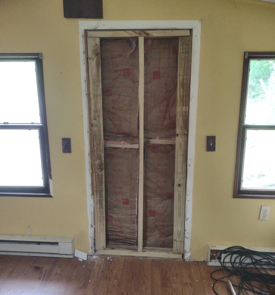 A tall doorway opening in a yellow wall showing bare studs and insulation, with a window on each side and a wood floor.