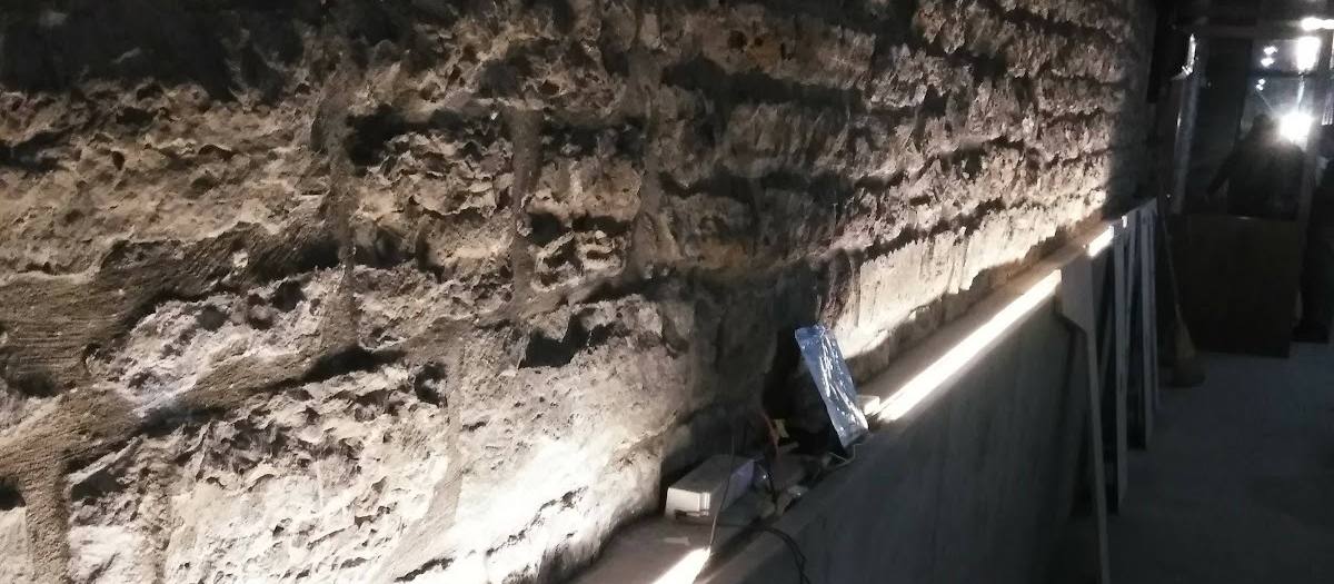 A strip of bright LED lighting running along the top edge of a concrete ledge at the foot of a rough stone wall, in a dim corridor.
